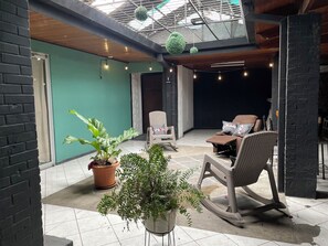 Un patio couvert aménagé avec des meubles en osier, des plantes en pot et une vue sur l’intérieur grâce à un plafond en verre.