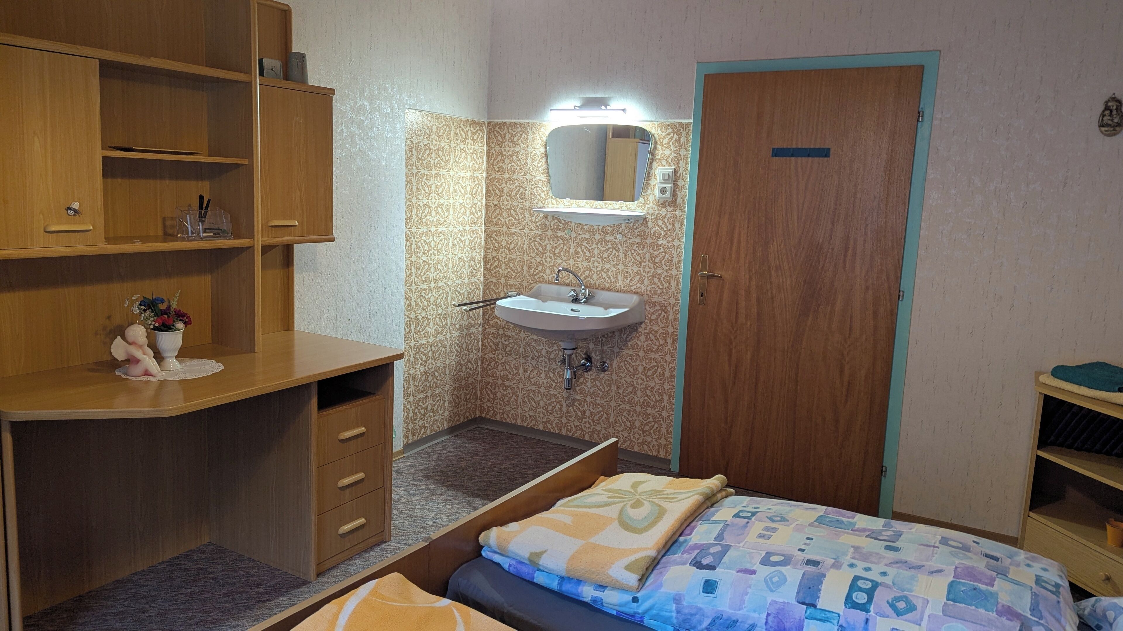 A small, compact bedroom with a wooden desk, a bedside table, a single bed with a patterned blanket, a built-in sink with a mirror, and a wooden wardrobe.
