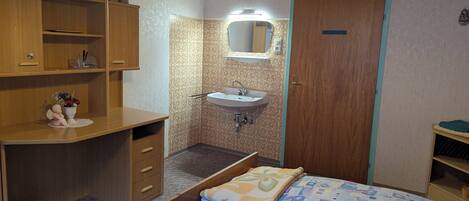 A small, compact bedroom with a wooden desk, a bedside table, a single bed with a patterned blanket, a built-in sink with a mirror, and a wooden wardrobe.