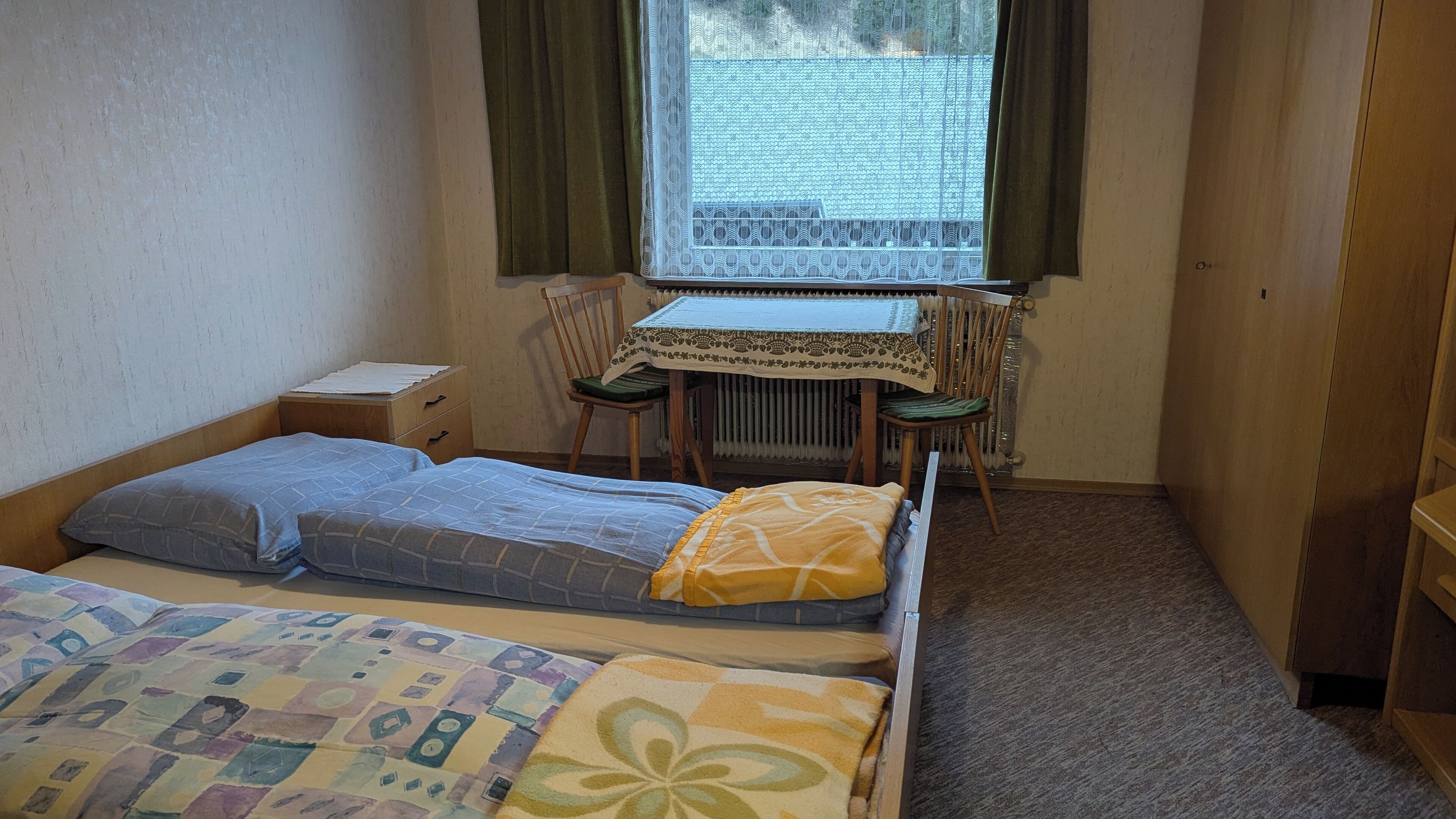 A small, compact room with two single beds, a wooden nightstand, a small table with a patterned tablecloth, a chair, and a radiator.