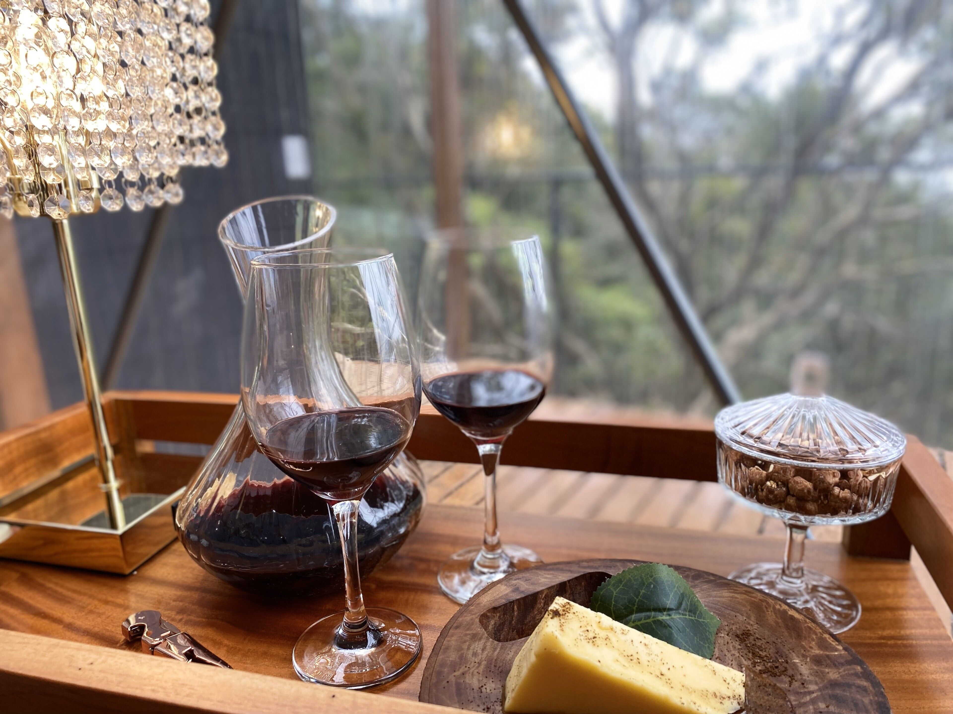 A wooden tray with two wine glasses, a pitcher, a cheese plate, and a coffee container.