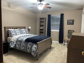 Une chambre à coucher avec un lit, un ventilateur de plafond, une fenêtre avec des rideaux, une chaise et une bibliothèque.