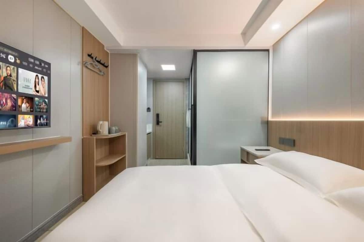 A modern hotel room with a large bed, a flat-screen TV mounted on the wall, a wooden shelf, and a sliding door leading to a bathroom.