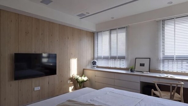 Une chambre à coucher avec un lit, un téléviseur fixé au mur, un bureau avec un ordinateur et une fenêtre avec des stores.