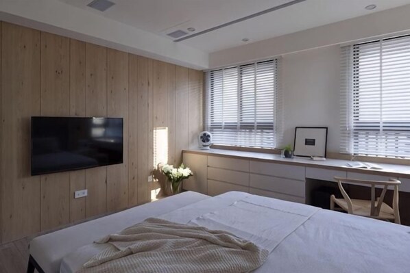 Un dormitorio con cama, televisor montado en la pared, escritorio con computadora y ventana con persianas.