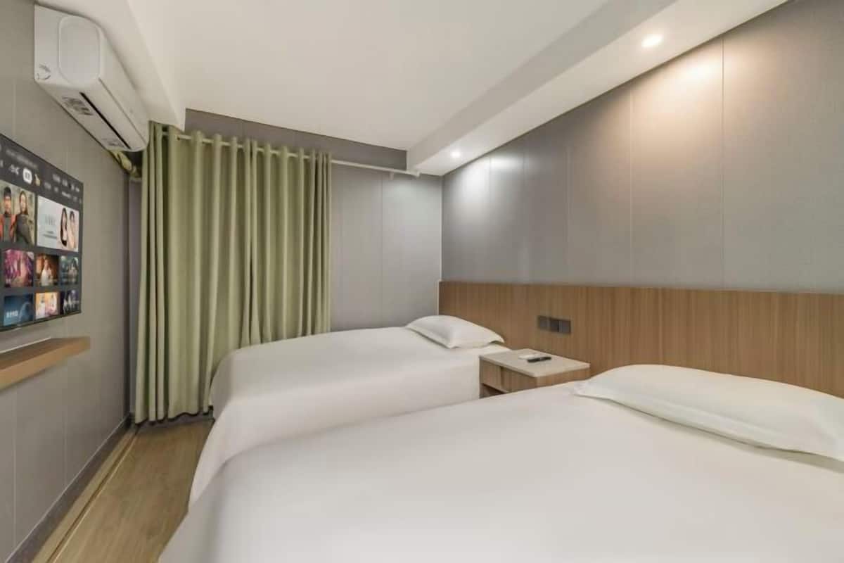 A hotel room with two beds, a nightstand, a wall-mounted air conditioning unit, and a curtain.