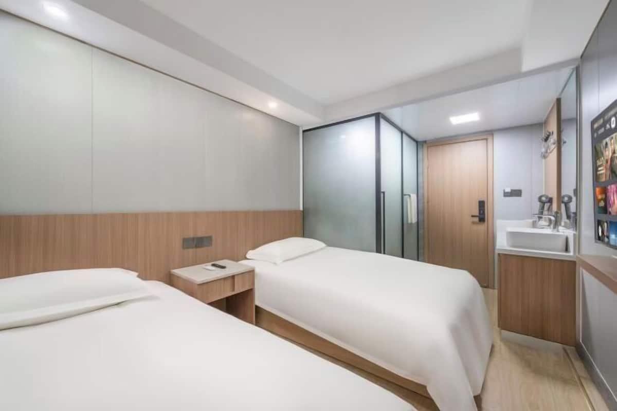 A hotel room with two beds, a wooden headboard, a glass partition, a wooden wardrobe, and a small bathroom with a sink and mirror.