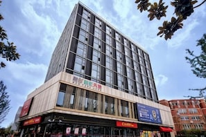 Un edificio de varios pisos con fachada moderna de vidrio, una zona comercial en la planta baja con señalización, y edificios residenciales adyacentes.