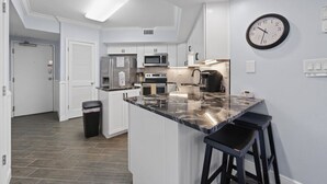 A modern kitchen with white cabinetry, a marble countertop, and a clock on the wall. - White Caps 102 with Pools (Orange Beach)