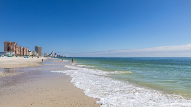 A beach with a clear sky, a sandy shore, and buildings in the background. - White Caps 102 with Pools (Orange Beach)