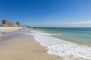 A beach with a clear sky, a sandy shore, and buildings in the background.