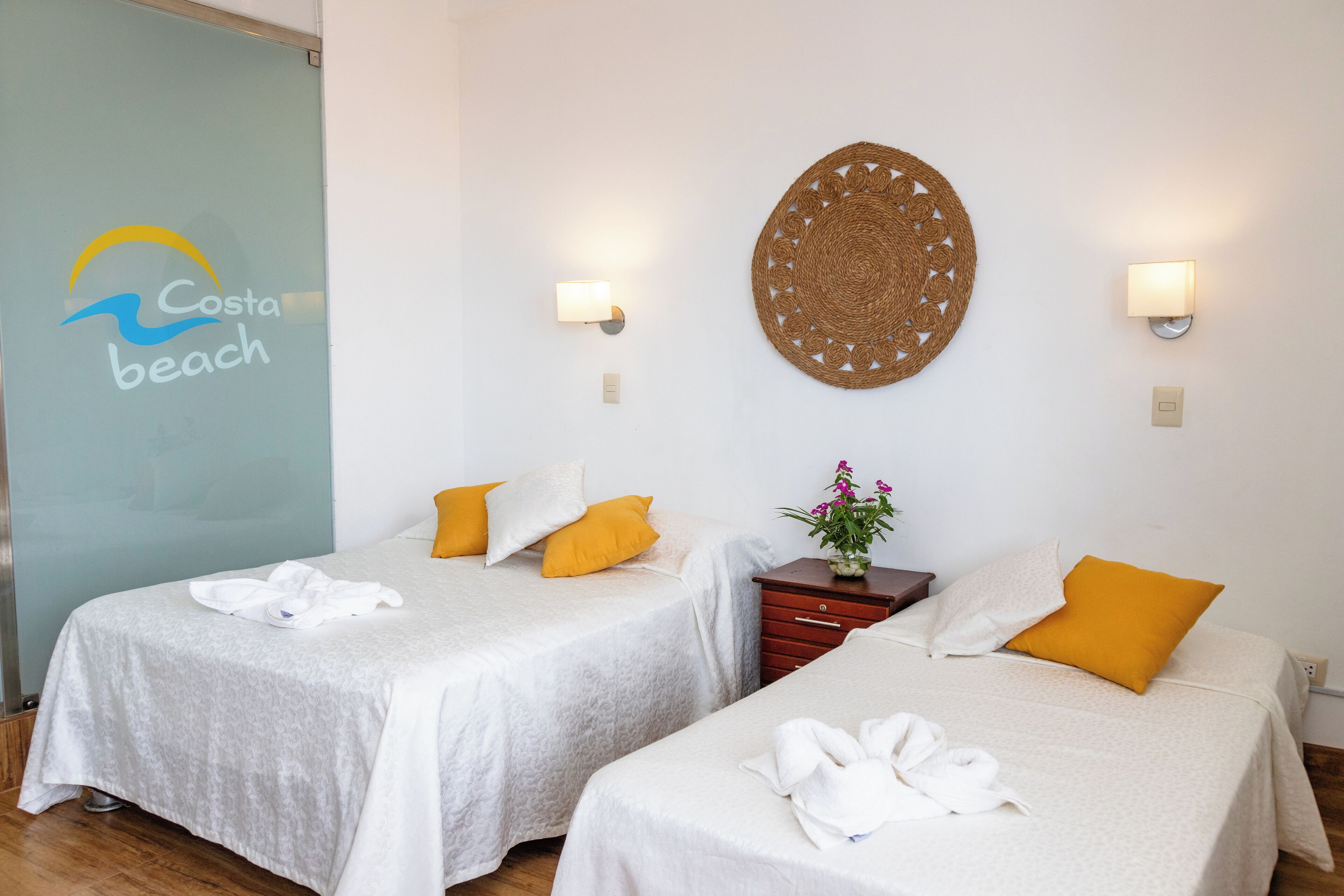 A hotel room with two beds, each with white linens and a single yellow pillow. There is a woven wall decoration above the beds and a small wooden nightstand with a vase of flowers.