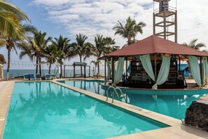 Une piscine dotée de chaises longues et d’un kiosque, entourée de palmiers et offrant une vue sur la mer.