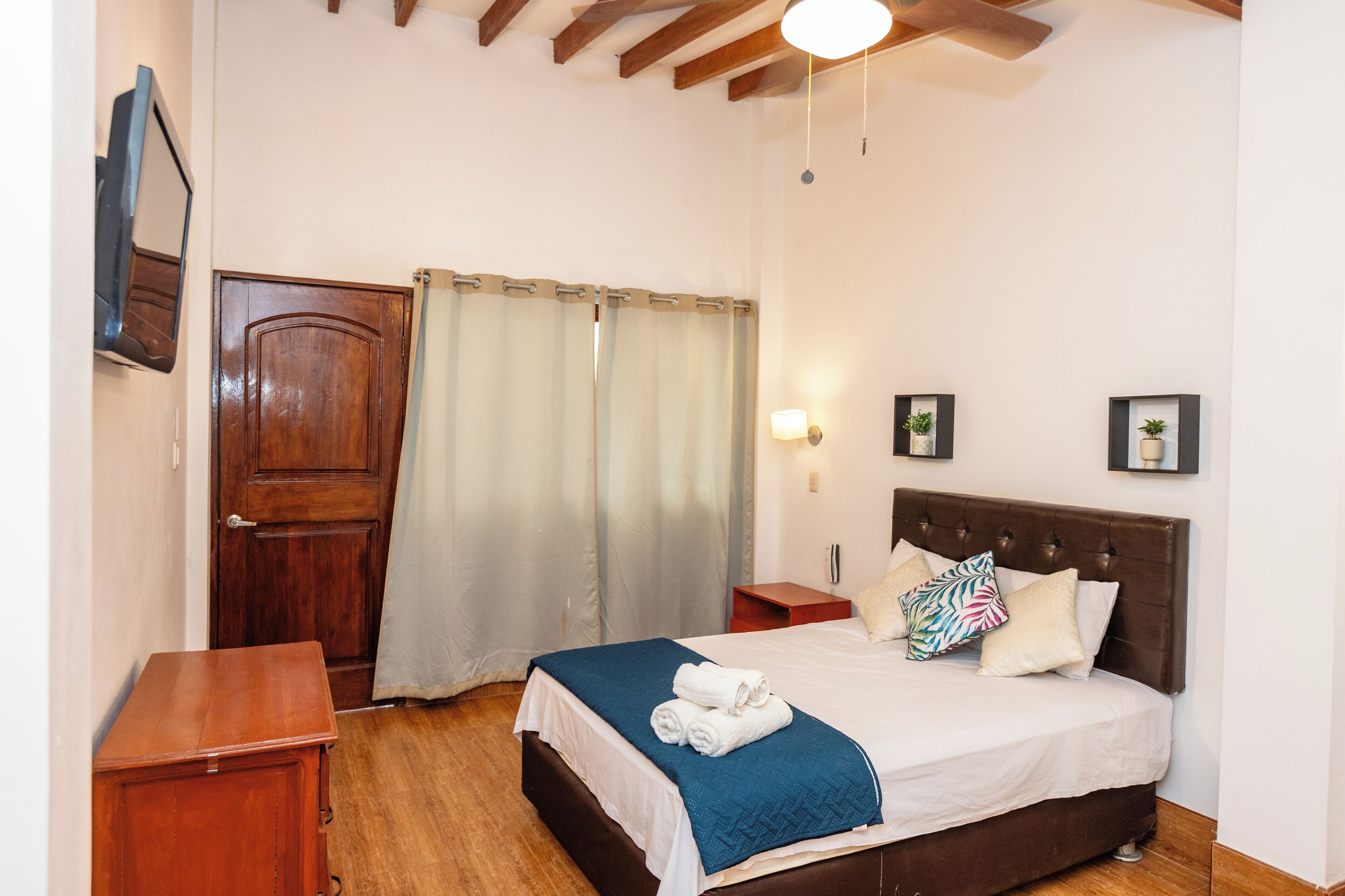 A bedroom with a wooden door, a bed with a blue blanket, a bedside table, and a nightstand with a lamp.