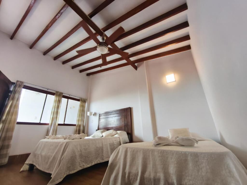 Habitación de hotel con dos camas, vigas de madera en el techo, una ventana con cortinas y una lámpara fijada a la pared.