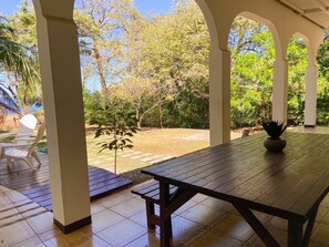 Eine überdachte Terrasse mit einem Holztisch und Stühlen, umgeben von einem Garten mit Bäumen und Rasen.