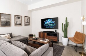 A modern living room with a flat-screen TV, a sofa, a coffee table, a cactus plant, and a vintage chair.