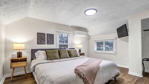 A bedroom with a large bed, two bedside tables, a flat-screen TV mounted on the wall, and a ceiling light.