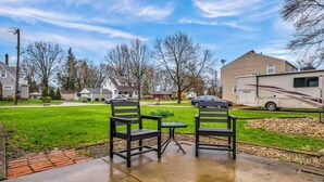 A residential area with two black chairs and a small table on a concrete patio, a beige RV in the background, and houses with lawns and trees.