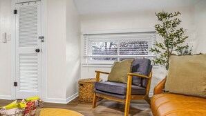 A living room with a wooden floor, a leather sofa, a wooden chair with cushions, a wicker basket, and a window with blinds.