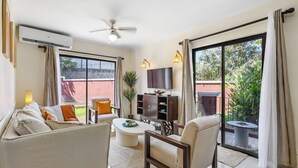 A modern living room with a sofa, armchair, coffee table, and TV. There is a balcony with plants and a view of greenery. - Tamarindo Escape: Exquisite One-Bedroom Condo (Tamarindo)