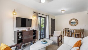 A living room with a flat-screen TV, a white sofa, a coffee table, and a dining area with wicker chairs. - Tamarindo Escape: Exquisite One-Bedroom Condo (Tamarindo)