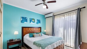 A bedroom with a wooden headboard, a bed with white bedding and a green throw, a nightstand with a lamp, and a ceiling fan. - Tamarindo Escape: Exquisite One-Bedroom Condo (Tamarindo)