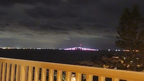 A night view from a balcony with a railing, overlooking a body of water and a distant cityscape with illuminated buildings.