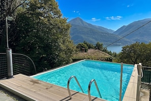 Ein Schwimmbecken mit Holzdeck und Metallgeländern, umgeben von Bäumen und Bergen im Hintergrund.