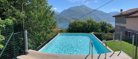 A rectangular swimming pool with a wooden deck and metal railings, surrounded by greenery and a mountainous backdrop.