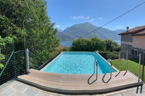 A rectangular swimming pool with a wooden deck and metal railings, surrounded by greenery and a mountainous backdrop.