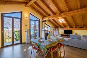 Ein Essbereich mit Tisch für vier Personen, einer Couch, einem Fernseher und einem Balkon mit Blick auf die Berge.