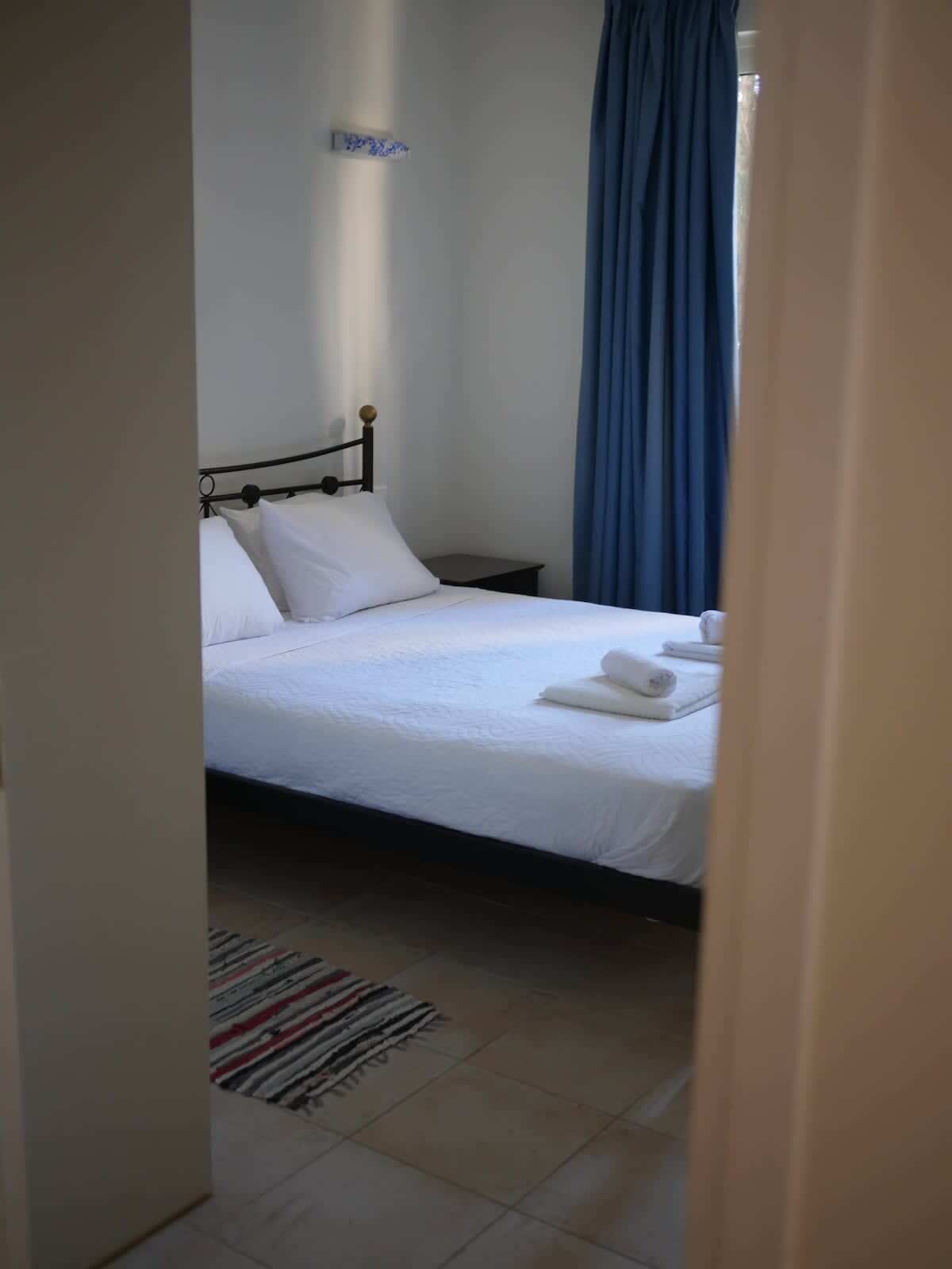Een opgemaakt bed met witte lakens, een klein nachtkastje en een blauw gordijn.