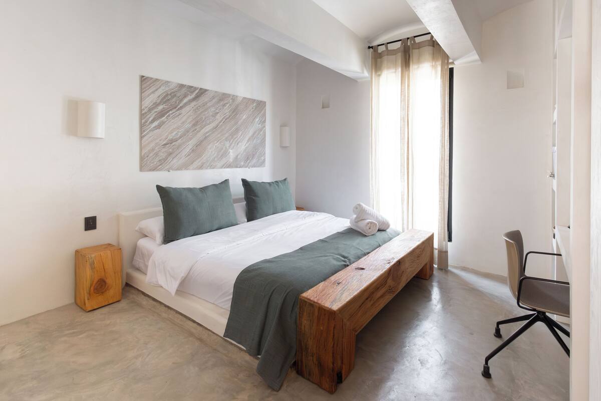 Un dormitorio moderno con una cama grande, un banco de madera, un escritorio y una obra de arte de mármol en la pared.