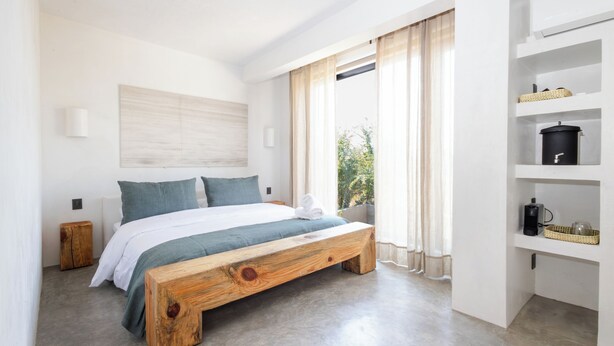 A modern bedroom with a wooden bed, white walls, and a large window with sheer curtains. - LePetit (Oaxaca)