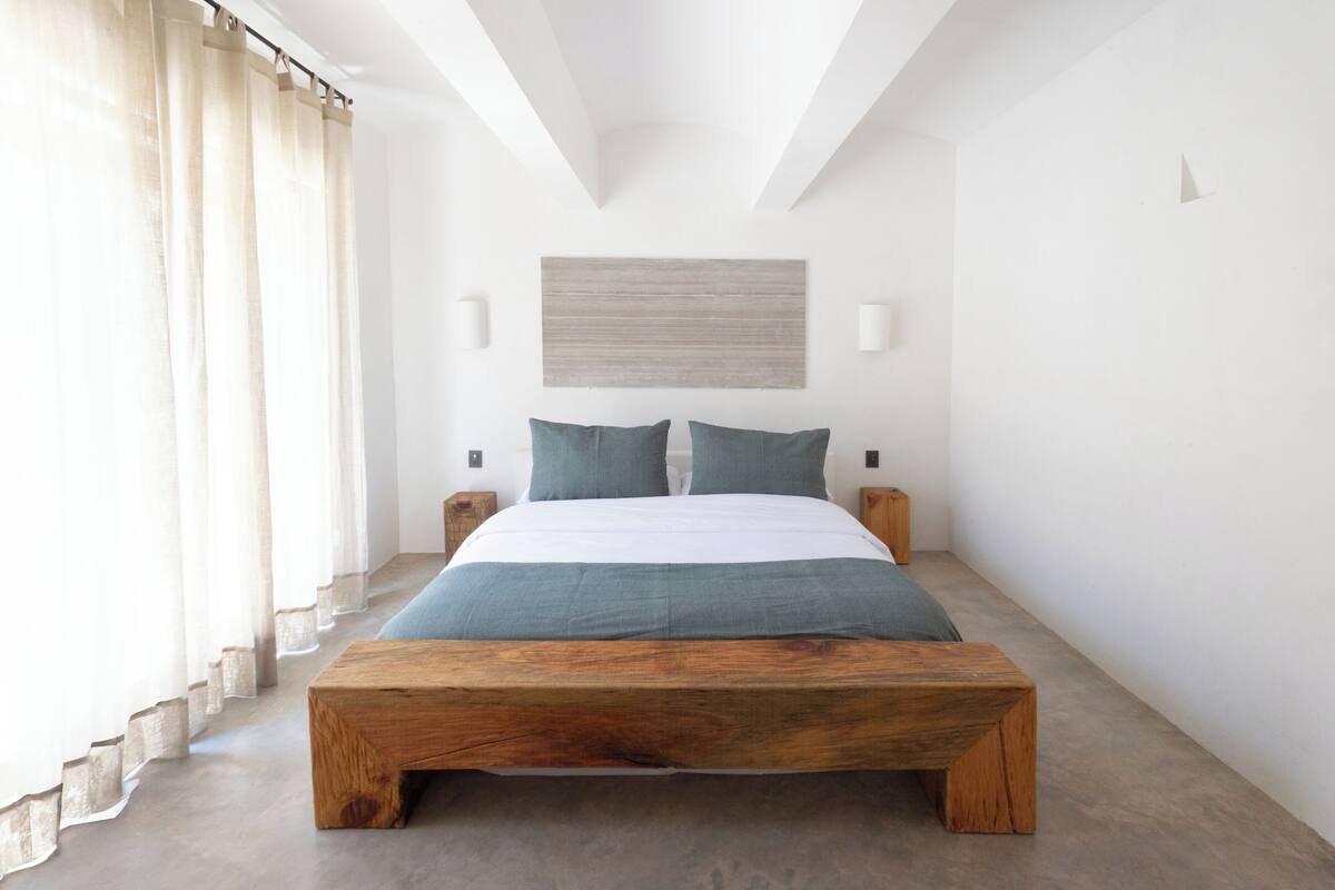 Un dormitorio moderno con una cama grande, un banco de madera, paredes blancas y un techo con vigas a la vista.
