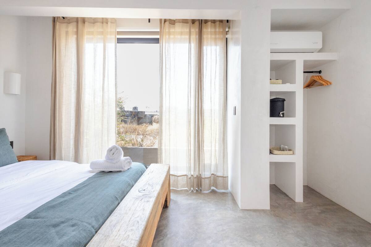 Un dormitorio con cama, un banco, una estantería con cestas y vistas al exterior.