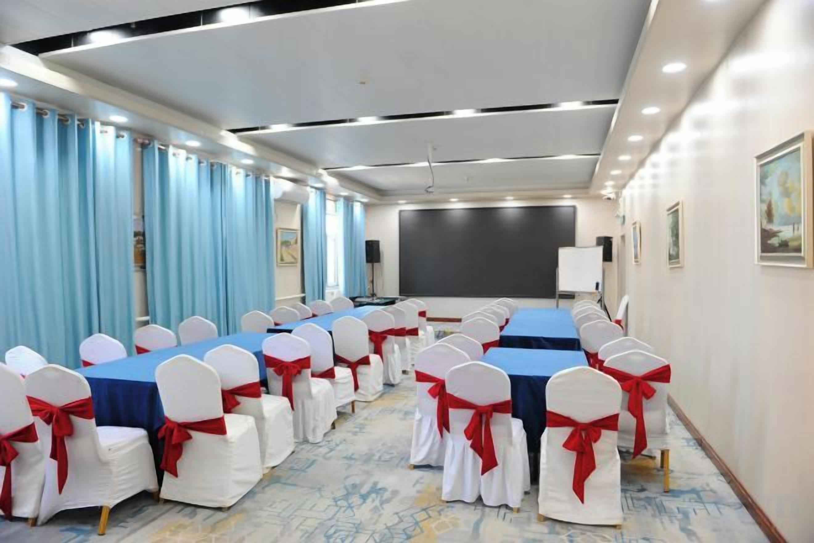 Un salón de conferencias con filas de mesas y sillas, una pantalla de proyector y una pizarra.