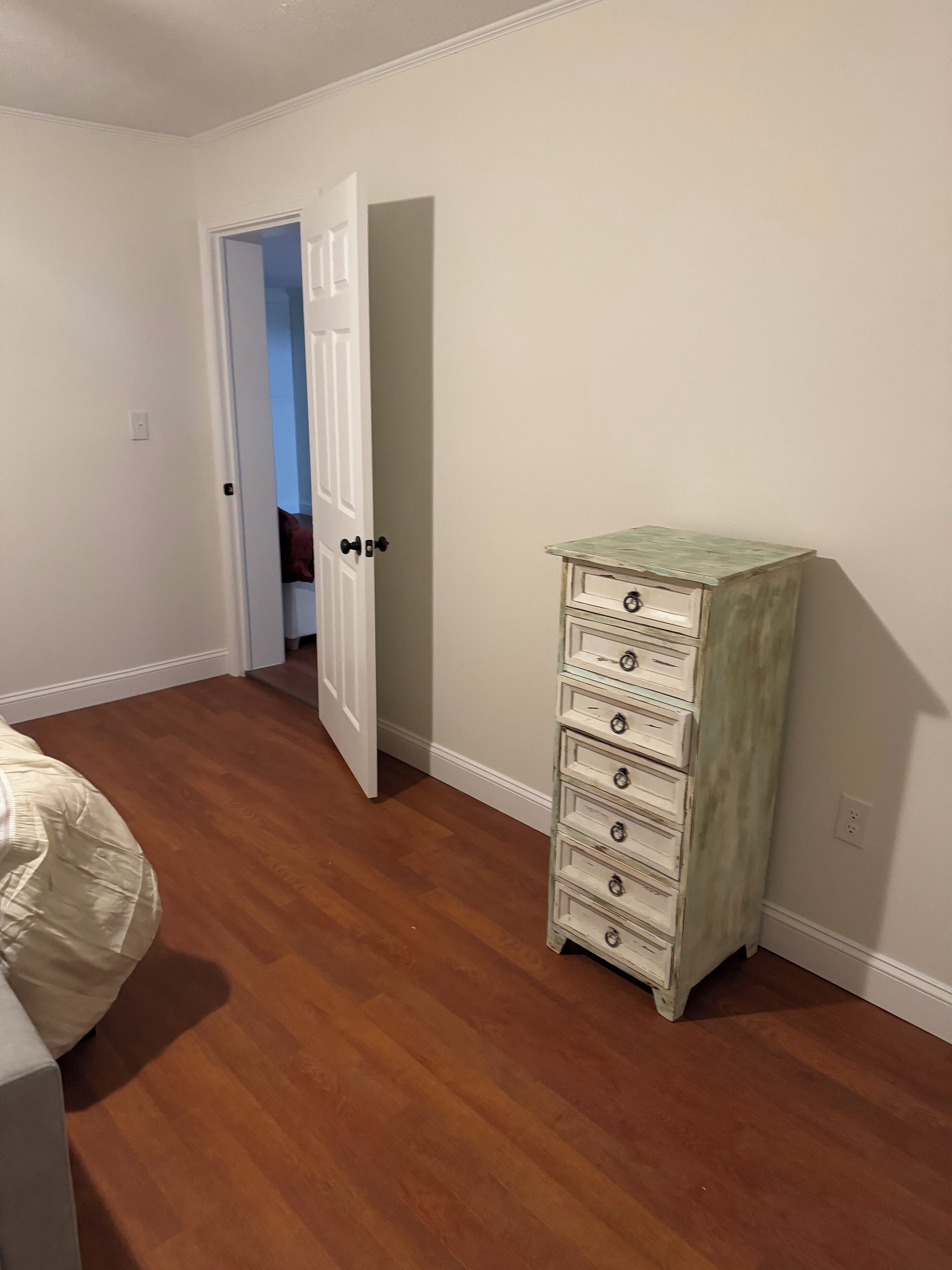 A bedroom with wooden flooring, a white door, and a tall, white dresser with drawers.