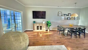 A dining area with a wooden table and chairs, a large window with blinds, a television mounted on the wall, and a cabinet with decorative items. - Spacious 8br/5.5ba home in Rehoboth Beach. WiFi, Pool, Pet friendly, Renovated. (Rehoboth Beach)