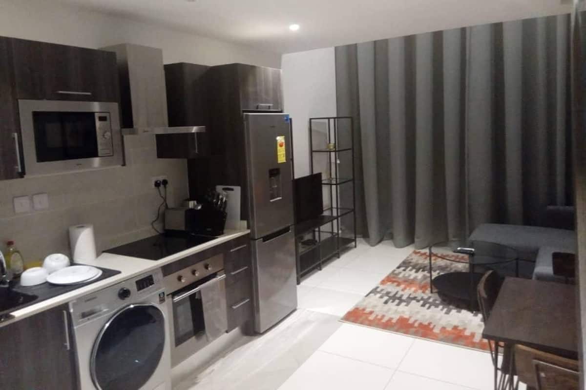 Een moderne keuken met roestvrijstalen apparatuur, een inbouwoven en een wasmachine. Er is een eethoek met een tafel en stoelen, en een zithoek met een bank en gordijnen.