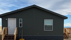 A small, dark-colored cabin with a white door and two windows, wooden stairs leading to the entrance, and a clear sky in the background.