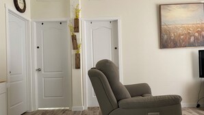 A room with a reclining chair, white doors, a clock, a painting, and a wooden floor.