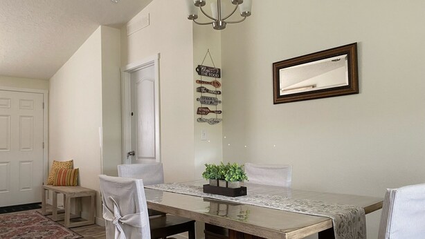 A dining area with a long table, chairs, a chandelier, a mirror, and a wall with a family-themed sign.