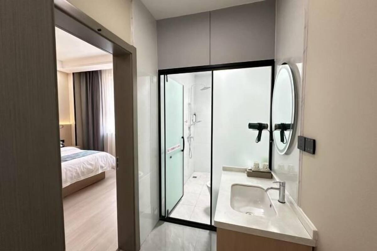 A modern hotel room with a glass-enclosed shower, a white sink, and a bed with a white duvet.