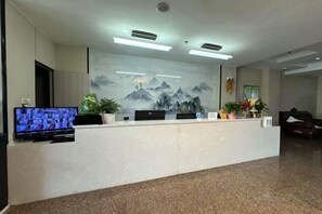 A reception desk with a flat-screen TV, potted plants, and a wall with a mountain painting. - Heshuo Huafeng Express Hotel (Hoxud)