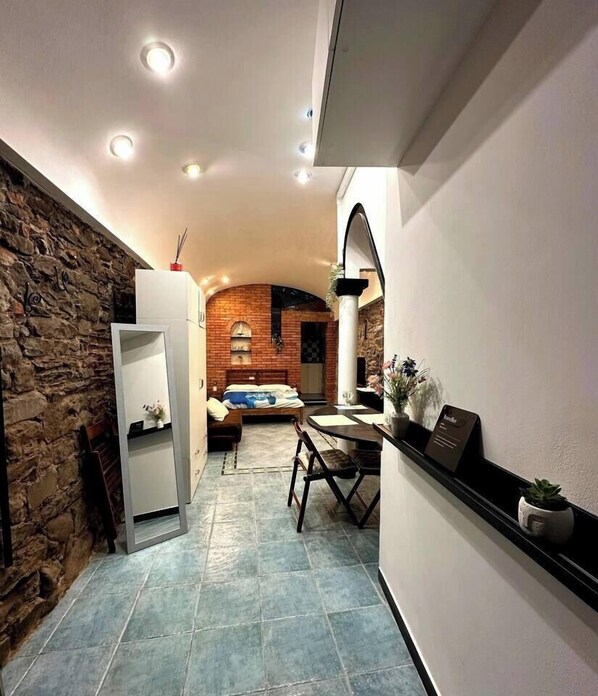 A narrow hallway with a stone wall on one side and a kitchen area on the other. A bed is visible in the background, and there is a small table with chairs in the kitchen area.
