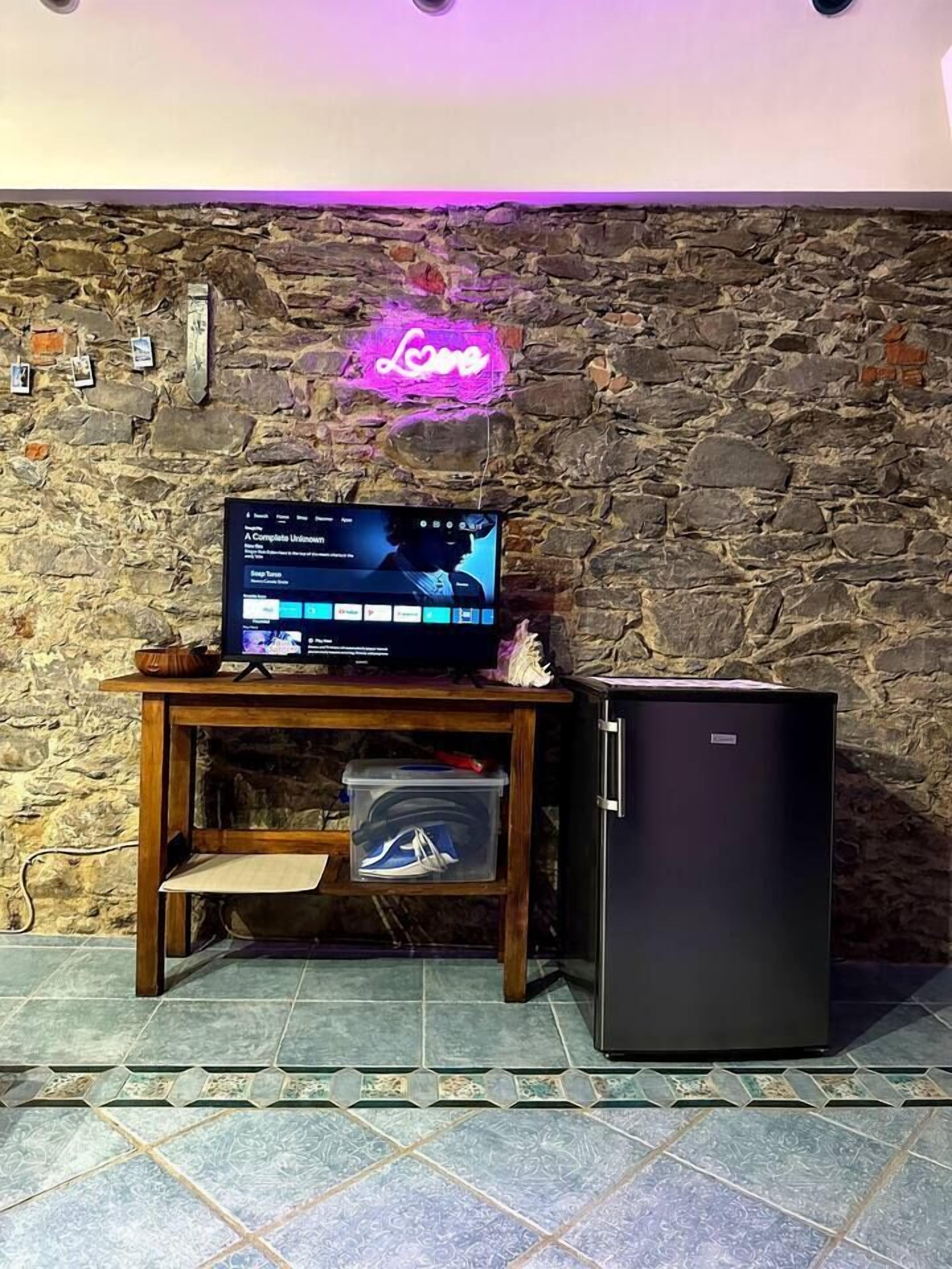 Ein Zimmer mit einer Steinwand, einem Neon-Schild mit der Aufschrift 'Love', einem Fernseher auf einem Holzständer und einem schwarzen Kühlschrank.