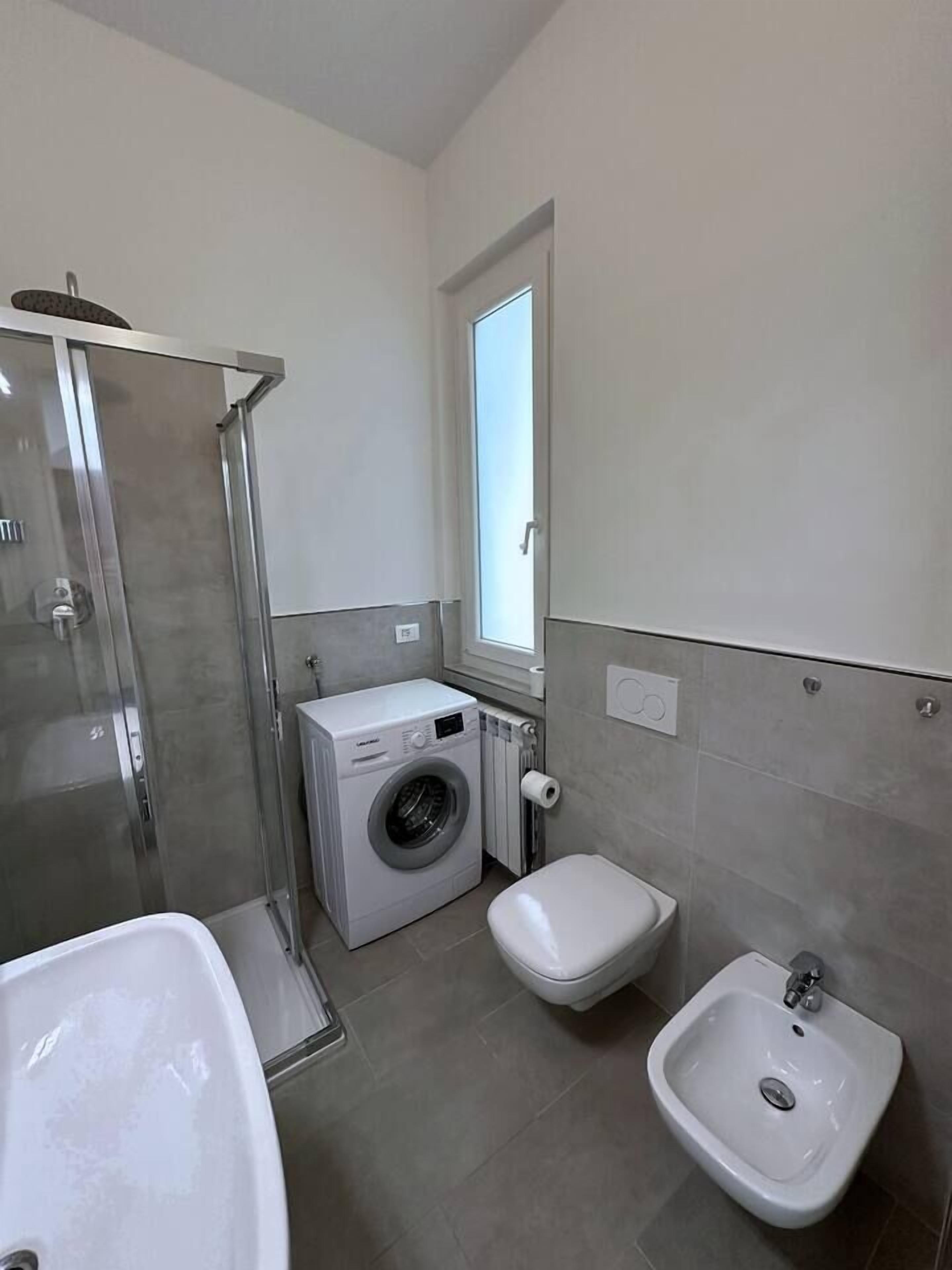 A compact bathroom with a glass-enclosed shower, a washing machine, a bidet, and a sink.