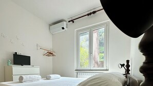 Ein Schlafzimmer mit Bett, Nachttisch, Lampe, Fernseher und einem Fenster mit Blick auf Grünflächen.